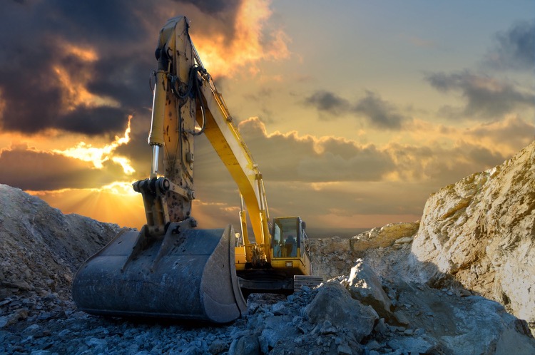 An excavator in a quarry. Sun beams cut through the clouds behind the machine.